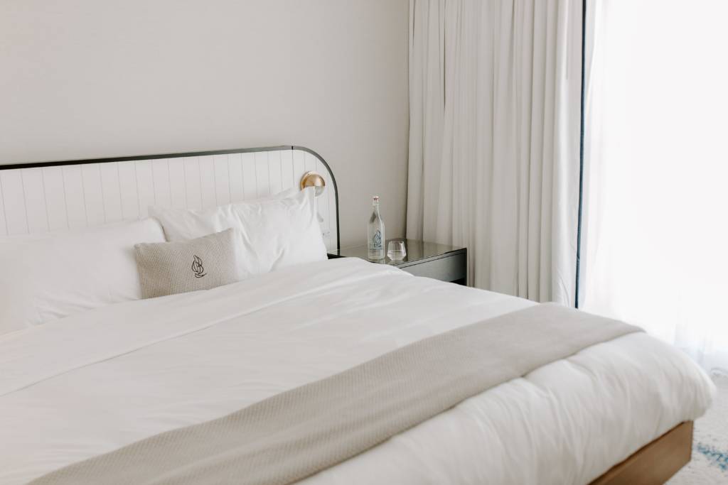 A bottle and glass of water on a nightstand next to a hotel bed