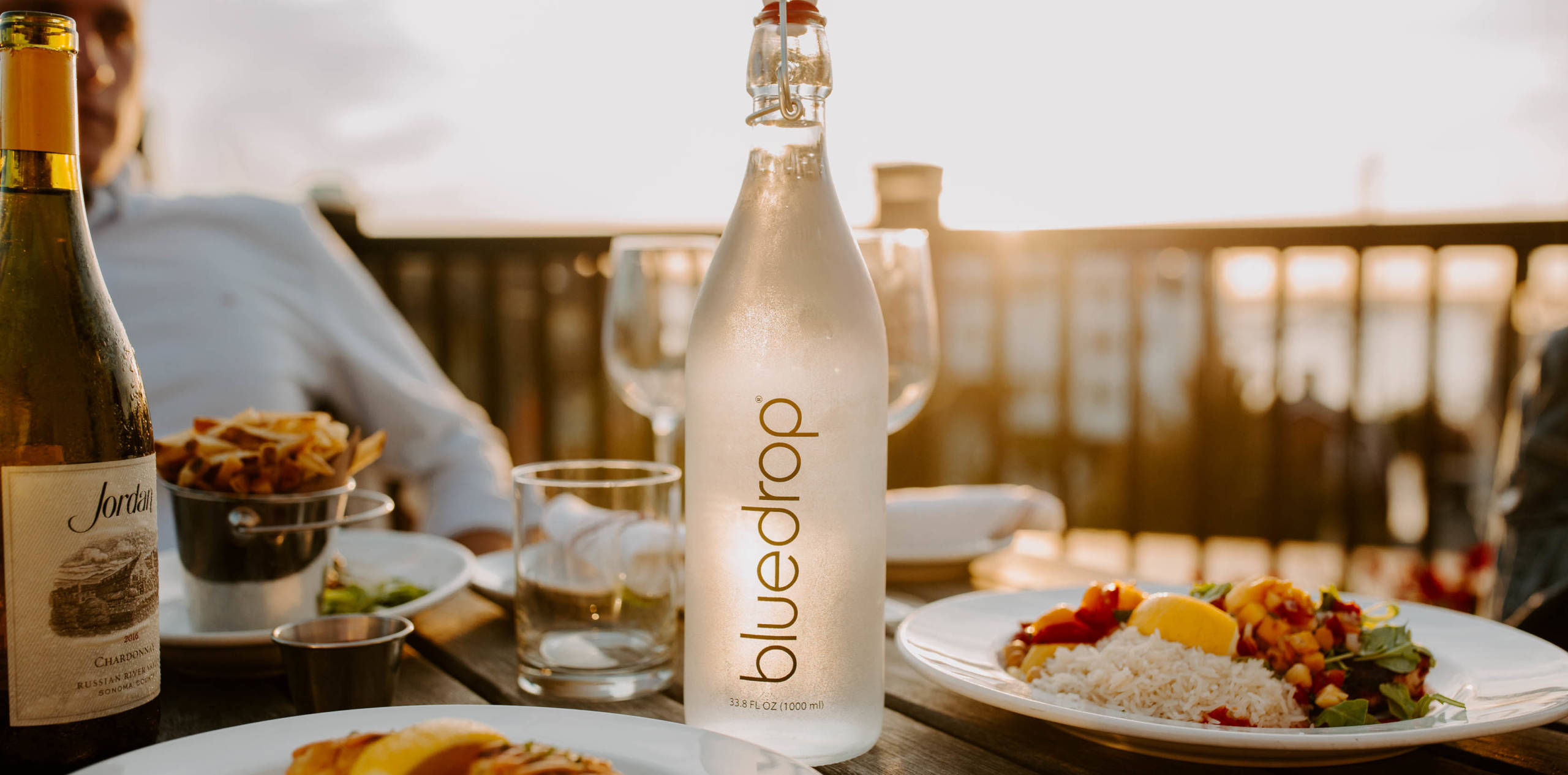 A bottle of bluedrop water sitting on a restaurant table with plates of food surrounding it.