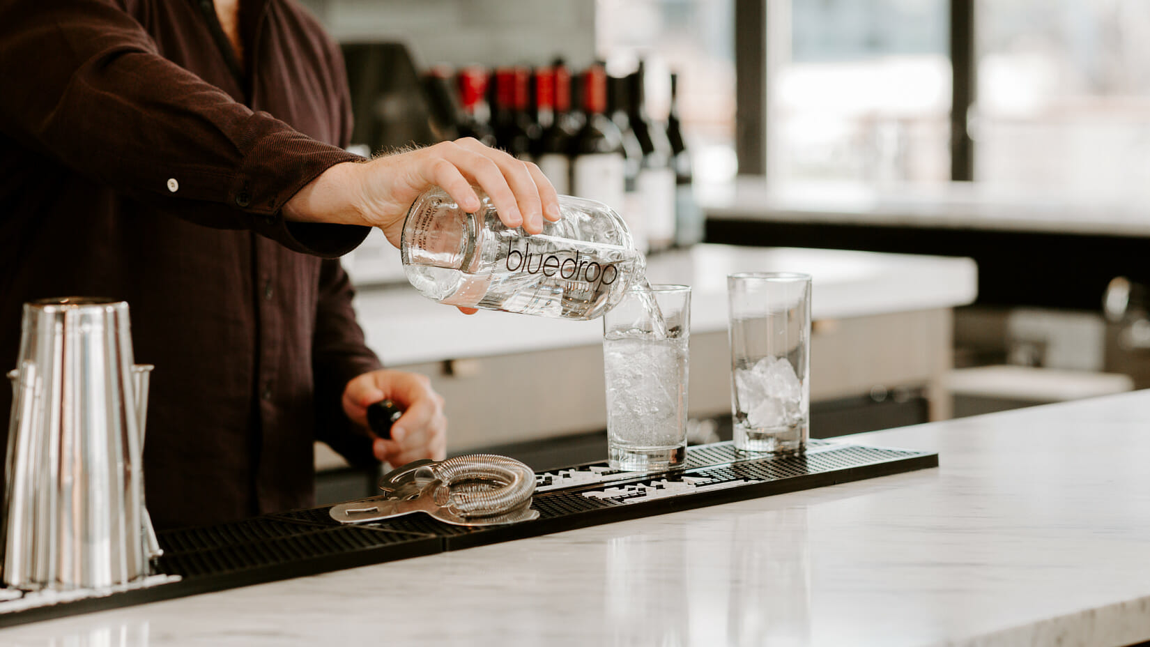 A person pouring water into a glass from a bluedrop water bottle at a restaurant bar.